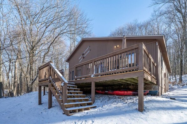 A wooden cabin with a staircase leading to a deck, surrounded by snow-covered ground and leafless trees.