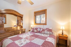 Quarto com cômoda de madeira, um espelho grande, janela com persianas e uma cama com colcha florida, além de abajures laterais.