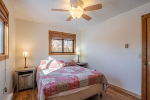 A bedroom with a bed, two bedside tables, a ceiling fan, a window with blinds, and a door.