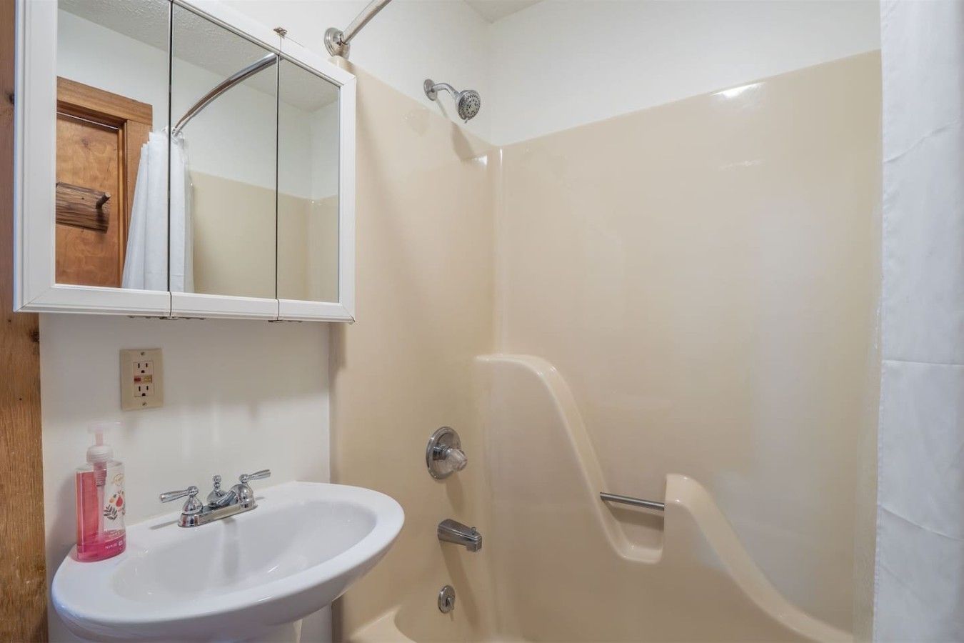 Un baño con lavamanos blanco, espejo, ducha con puerta de cristal y puerta de madera.