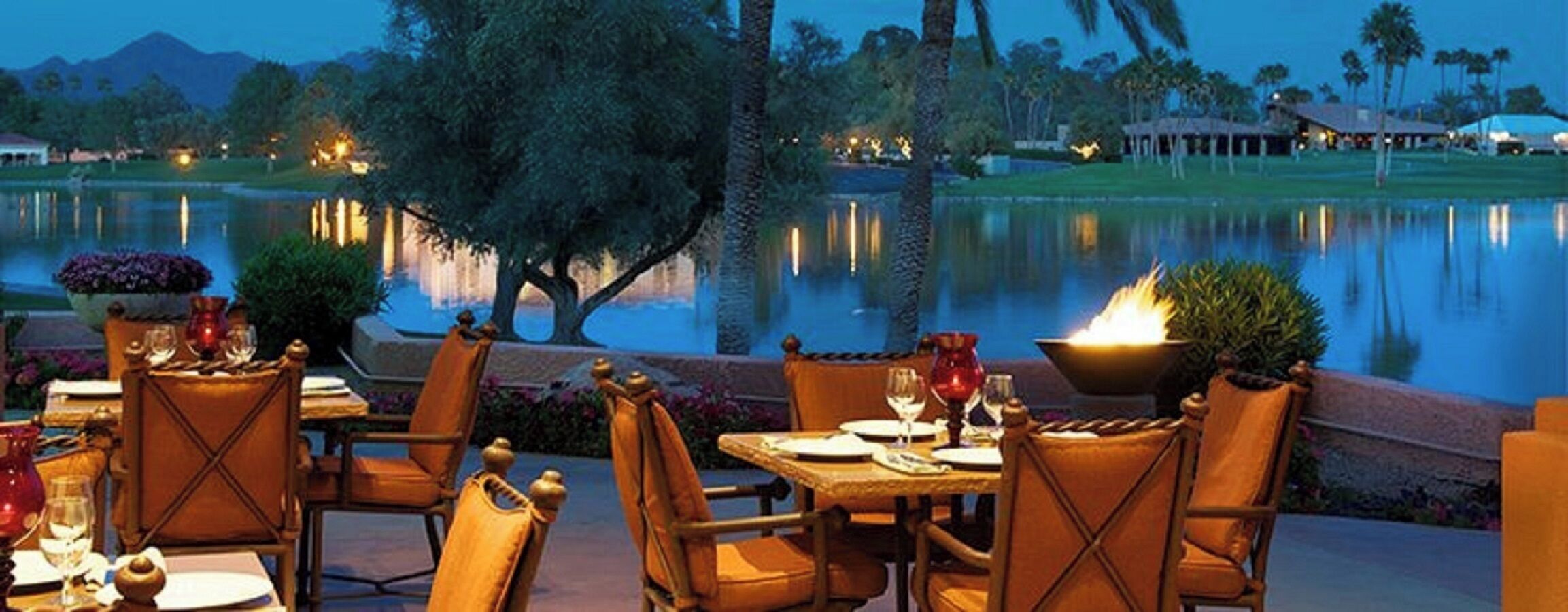 An outdoor dining area with tables and chairs, a fire feature, and a view of a body of water and distant buildings.