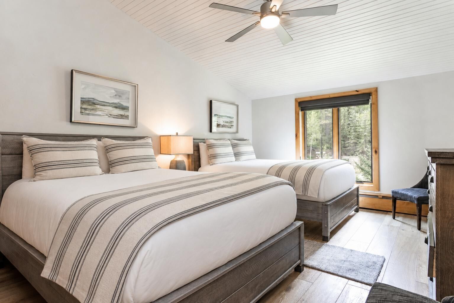 A bedroom with two beds, a ceiling fan, a window with a view of trees, and a wooden dresser.