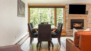 A dining area with a table and chairs, a fireplace, and a large window with a view of trees. - Riva Ridge Premium 745 | 2BR Vail Village (Vail)
