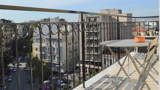 A balcony with a table and chairs, overlooking a city street with buildings and parked cars. - Vintage,spacious apartment in the heart of Athens! (Athina)