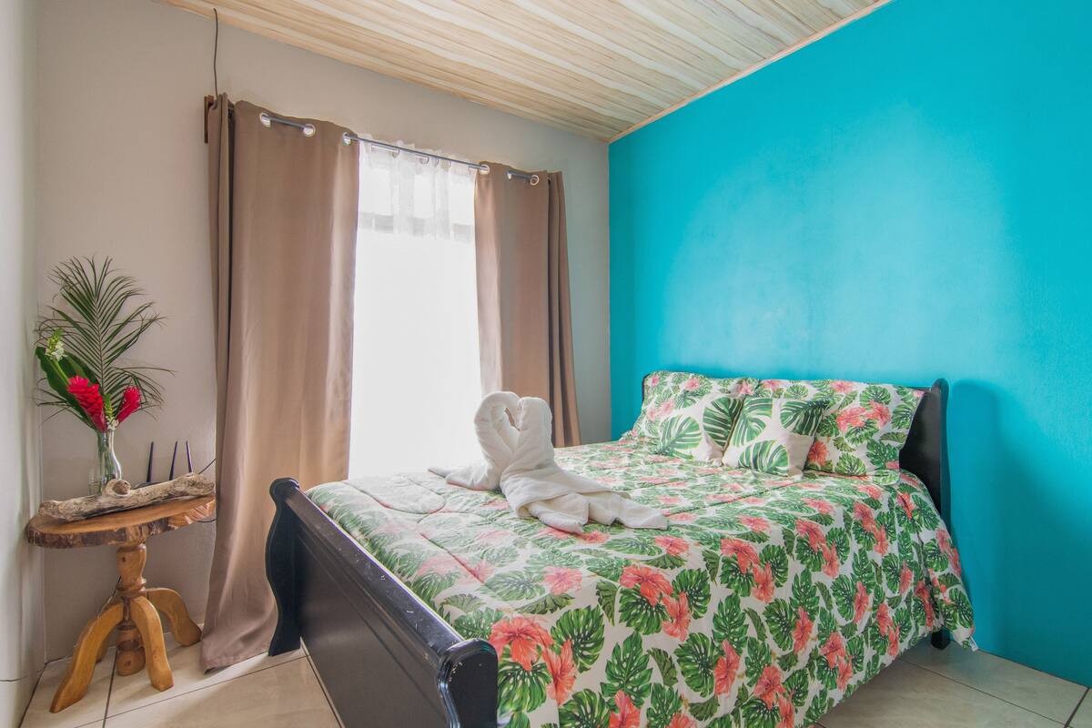 Une chambre à coucher avec un lit, une petite table sur laquelle est posé un vase, et une fenêtre avec des rideaux.