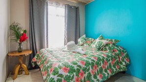 Une chambre à coucher avec un lit, une petite table en bois sur laquelle est posé un vase de fleurs, et une fenêtre avec des rideaux.