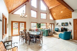 A spacious living room with a high ceiling, large windows, a dining area with a glass table, and a cozy seating area with a blue sofa.