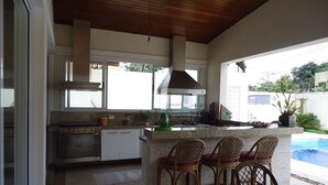 A modern kitchen with white cabinets, a stainless steel range, and a large window. There are three wicker bar stools and a small pool visible in the background. - SINGLE STOREY HOUSE - COMFORT AND LEISURE - ENSEADA - GUARUJÁ - (Guarujá)