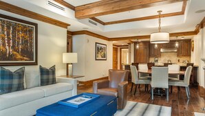 Una sala de estar con un sofá, un reposapiés azul, un comedor con sillas, una cocina con gabinetes de madera y un cuadro grande en la pared.