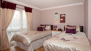 Ein Schlafzimmer mit zwei Betten, einem Fenster mit Vorhängen, einem Nachttisch mit Lampe und Wandbildern.