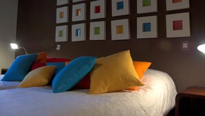 Un letto ordinato con cuscini colorati, una parete scura con cornici quadrate e un tavolino con una lampada.