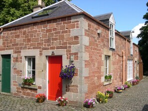 A stone building with a red door, multiple windows, and flower boxes on the ground. - Cottage Located In A Stunning South Facing Two Acre Private Garden Setting (Melrose)