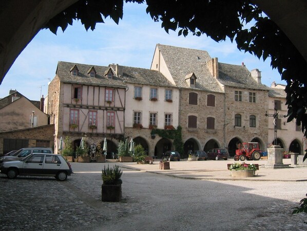 A traditional European town square with stone buildings, a cobblestone ground, and a red tractor in the background.
