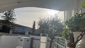 Um pátio ao ar livre banhado pelo sol, com cadeiras brancas, uma mesa pequena e plantas em vasos.