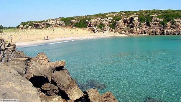 Uma praia costeira com águas turquesa cristalinas, afloramentos rochosos e uma faixa de areia.