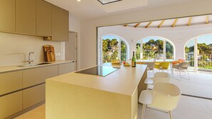 Una cucina moderna con isola centrale, zona pranzo con tavolo e sedie, e ampie finestre ad arco che si affacciano su vegetazione ed edifici.