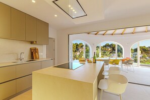 A modern kitchen with a central island, a dining area with a table and chairs, and large arched windows offering a view of greenery and buildings. - Villa Mar Alborán, Great Seaviews, 5-10 Min Easy Stroll Town & Beaches (Moraira)