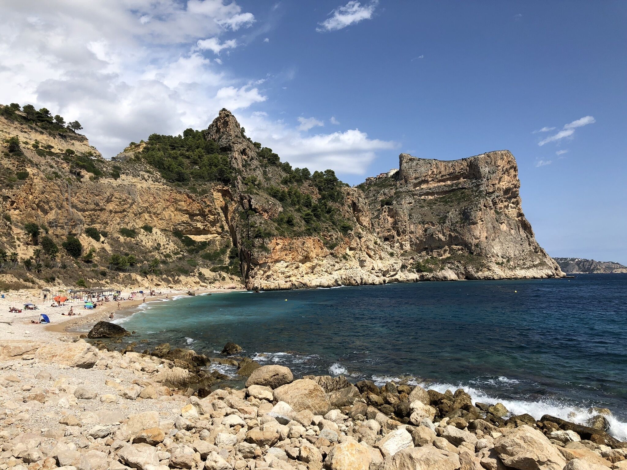 Una playa rocosa con aguas azules cristalinas, un acantilado con una cueva y algunas personas en la orilla.