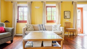 Uma sala de estar com um sofá, uma mesa de centro e uma poltrona. Há duas janelas com cortinas, um abajur e um quadro na parede.