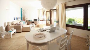 A modern living room with a white sofa, a dining table with chairs, a laptop, and a large window with curtains. - VILLA DEL MARE IN CHIA, WITH POOL OVERLOOKING THE SEA AND TOWER (Domus de Maria loc. Chia)