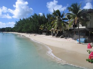 Une plage à l’eau turquoise et limpide, une rive sablonneuse et une rangée de bâtiments dotés de balcons donnant sur la plage.