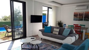 A modern living room with a grey sofa, a white coffee table, and a blue armchair. There is a TV mounted on the wall and a dining area with a table and chairs. - Romantic luxury "honeymoon" hideaway in the hills above Kalkan in a rural idyll (Kas)