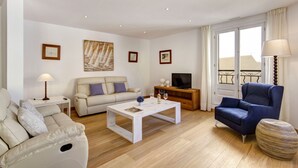 Ein Wohnzimmer mit Sofa, Sessel, Couchtisch, TV-Schrank und einem Fenster mit Vorhängen.