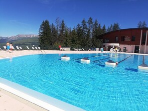 Ein Swimmingpool mit klarem, blauem Wasser, umgeben von Liegestühlen und einem Gebäude mit der Aufschrift 'PISCINE'.