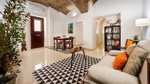 A modern living room with a geometric patterned rug, a sofa with orange cushions, a bookshelf, and a dining area with a table and chairs. - Casa Becco dos Assucares - free GARAGE - Historic Center (Évora)