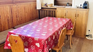 A cozy dining area with a wooden table covered in a red tablecloth, wooden chairs, and a wooden ceiling.