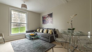 A modern living room with a grey sofa, a glass coffee table, a glass dining table with chairs, a large window with a view of a garden, and a painting on the wall. - Central Windsor Apartment Facing The Castle (Windsor)
