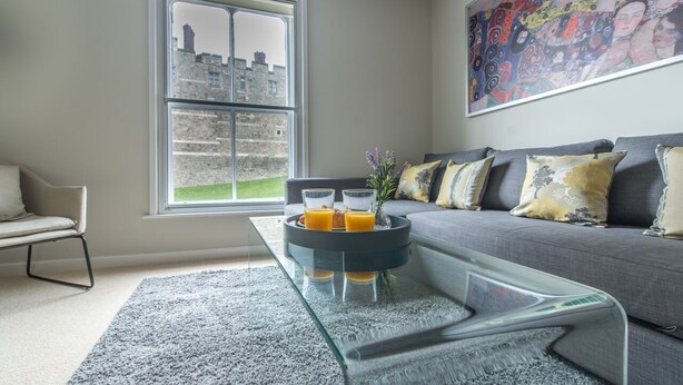 A modern living room with a grey sofa, a glass coffee table with orange juice, and a large window with a view of stone buildings. - Central Windsor Apartment Facing The Castle (Windsor)