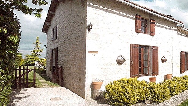 A white building with brown shutters, a gravel driveway, and a small garden with yellow flowers.