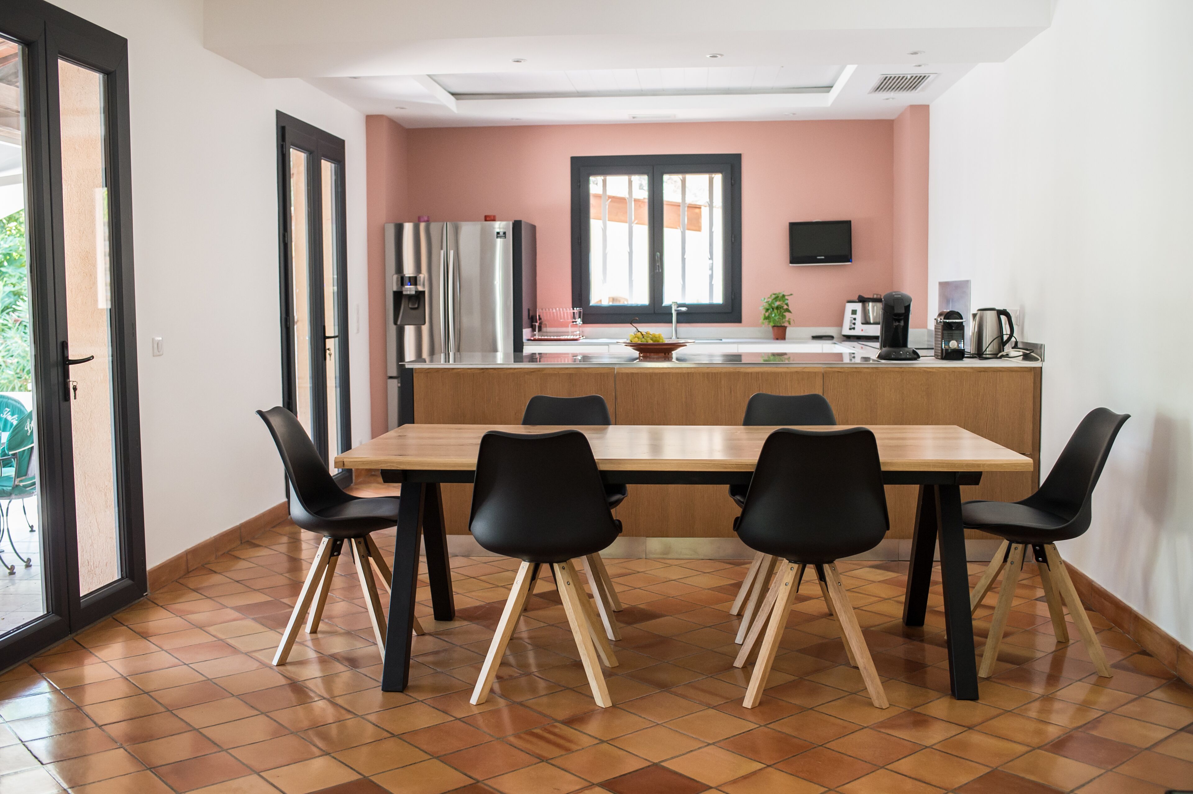 Une cuisine moderne avec une table à manger en bois et des chaises noires, un réfrigérateur et une fenêtre avec des stores.