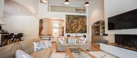 A spacious living room with a high ceiling, a large painting, a flat-screen TV, and a kitchen in the background.