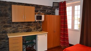 Un dormitorio con cama, escritorio con silla, refrigerador pequeño, microondas y televisor sobre una pared de piedra.