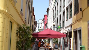 Una calle estrecha con mesas al aire libre bajo un paraguas rojo, un bar de tapas y un bote de basura verde.