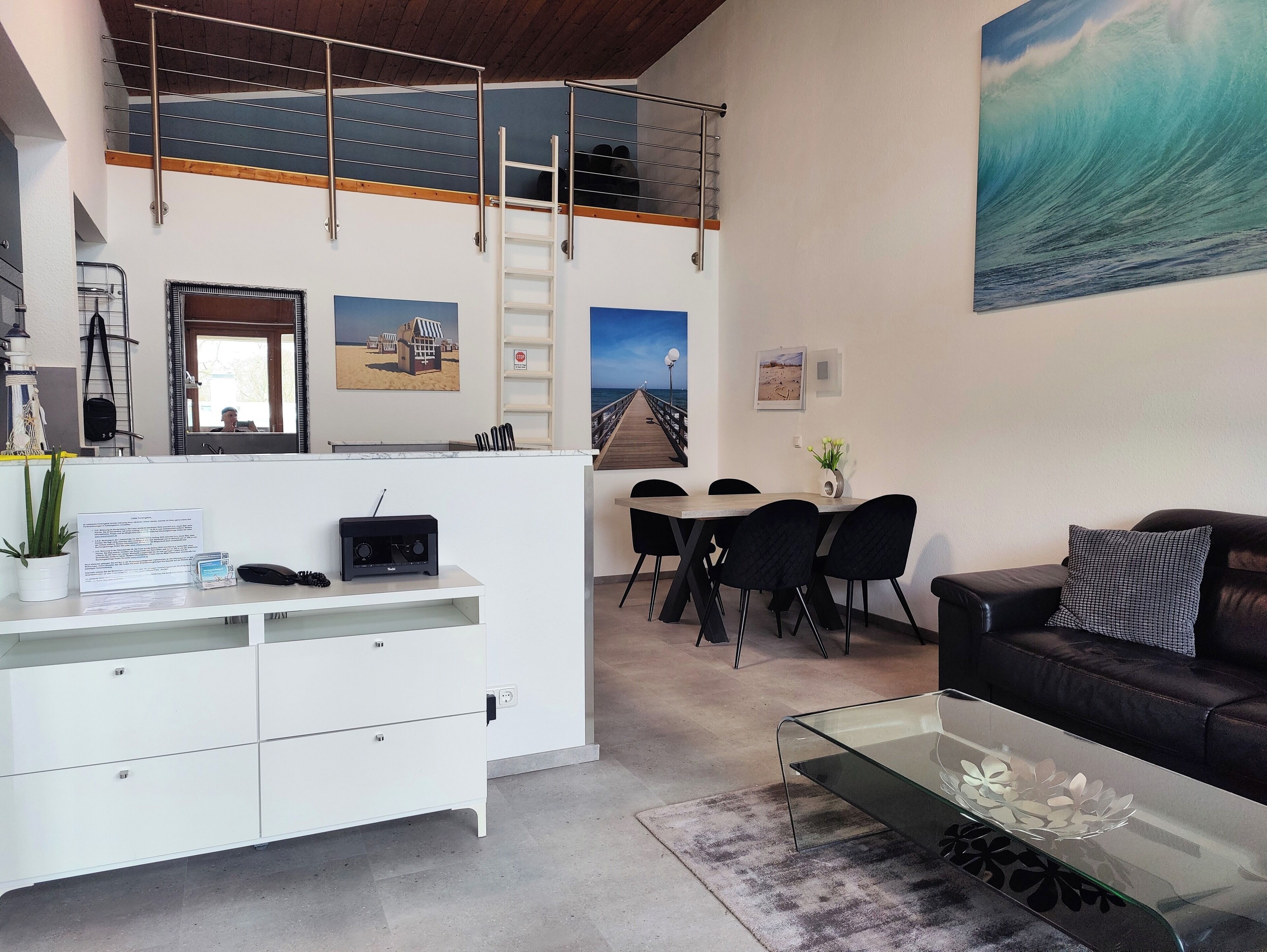 A modern living room with a loft bedroom above, featuring a balcony, a dining area, and a large beach-themed painting.