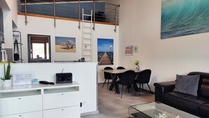 A modern living room with a loft bedroom above, featuring a balcony, a dining area, and a large beach-themed painting. - Attic apartment, Strandstr., side view. Sea view, 150 m to the Baltic Sea, renovated in 2025 (Kühlungsborn)