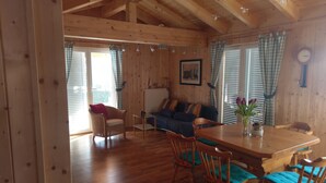 A wooden cabin interior with a dining table, chairs, a sofa, and a window with curtains.