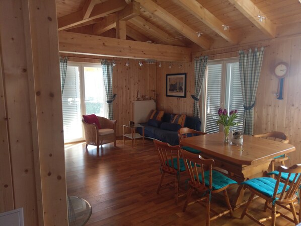 Interior de una cabaña de madera con una mesa de comedor, sillas, un sofá y una ventana con cortinas.