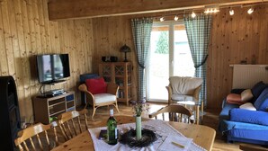 A cozy wooden cabin interior with a television, wicker furniture, a dining table with a lace tablecloth, and a view through large windows.