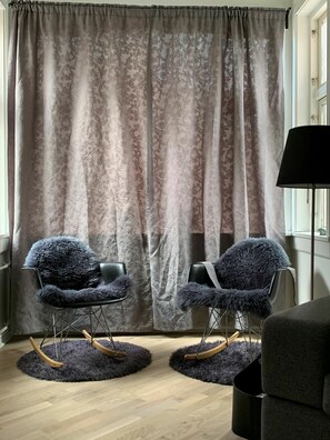 Een kamer met een groot raam, twee ruime fauteuils met zachte kussens, een zwarte vloerlamp en een kamerkleed met een patroon.