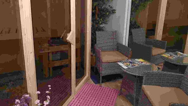 A room with a wooden door, a small table with a chair, a wicker chair, and a small table with magazines.