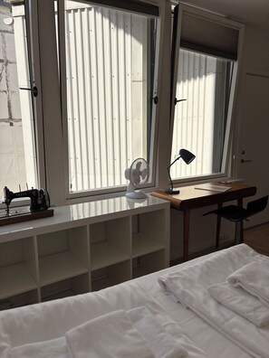 A bedroom with a bed, a desk, a sewing machine, and a window with blinds.