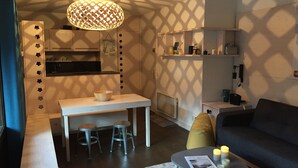 Uma sala de estar moderna com um sofá, uma mesa de centro com velas, uma área de refeições com banquinhos e uma cozinha ao fundo.