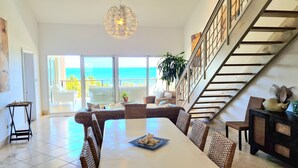A spacious living area with a dining table, wicker furniture, and a view of the ocean through large sliding doors.
