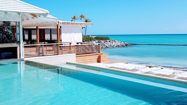 A clear blue pool with a view of the ocean, a covered outdoor seating area, and a building with a sign that reads "BEACH".