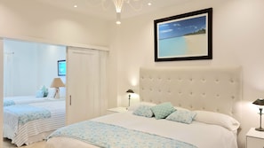 Um quarto com uma cama grande, uma mesa de cabeceira, uma luminária, um quadro com uma praia e uma porta para outro cômodo.