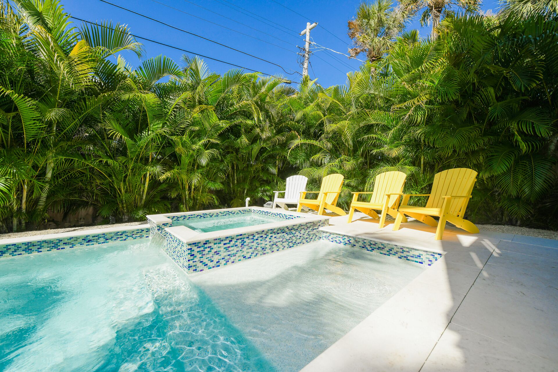 A swimming pool with tiled edges, surrounded by lush green foliage and yellow Adirondack chairs.